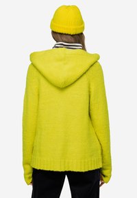 Jasně žlutá pletená mikina s žebrovaným lemem a manžetami, s kapucí a hladkým, jemným materiálem. Nosená s pruhovaným límcem pod ní.