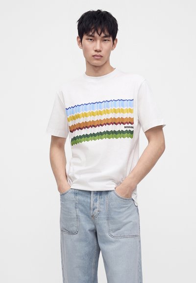Νέος άνδρας με λευκό t-shirt με πολύχρωμες ζικ-ζακ ρίγες και ανοιχτό μπλε τζιν, όρθιος με τα χέρια στις τσέπες μπροστά σε απλό φόντο.