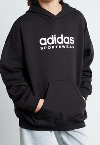 Musta huppari, valmistettu puuvillasekoitteesta, jossa edessä kengurutasku ja valkoinen "adidas sportswear" -logo rinnassa.
