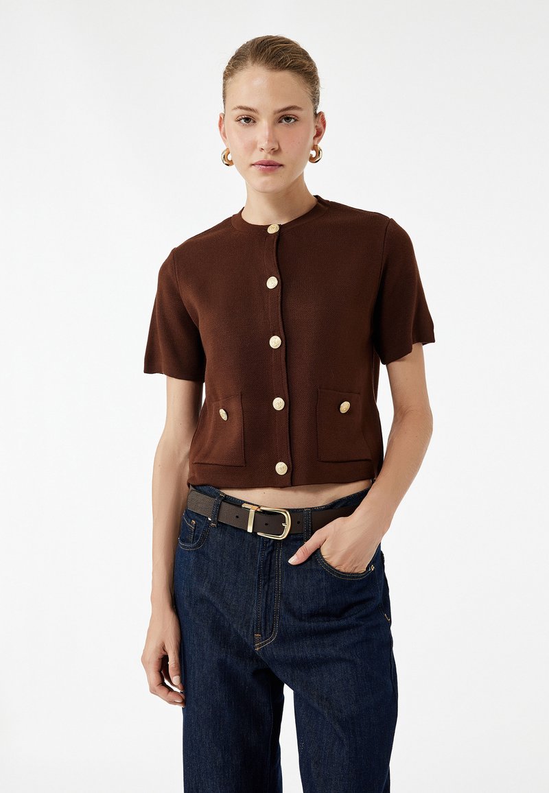 Braune, kurzärmelige, cropped Strickjacke aus geripptem Stoff mit goldenen Knöpfen und zwei vorderen Taschen. Getragen über dunkellblauen Jeans und einem schwarzen Gürtel.