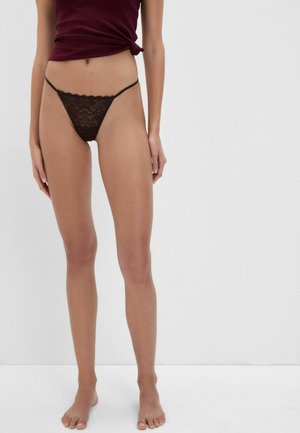 Femme portant un string en dentelle foncé et un haut bordeaux noué, debout pieds nus sur un fond clair uni.
