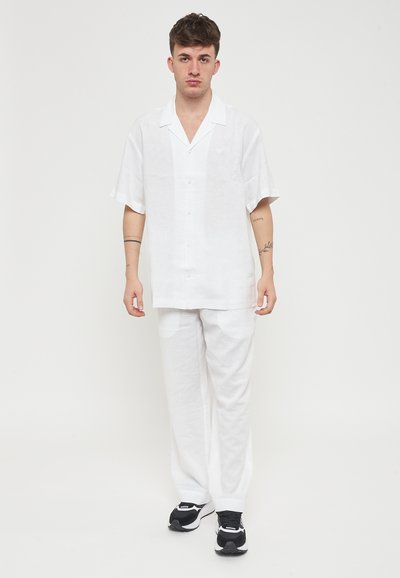 Camicetta bianca in lino a maniche corte con chiusura a bottoni, abbinata a pantaloni bianchi in lino coordinati. Sneakers nere con accenti bianchi e grigi.