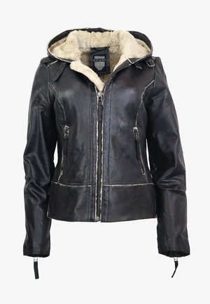 Kvinde iført en sort læderjakke med beige lammeskindsfôr, sort rullekrave, tætsiddende sorte bukser og sorte ankelstøvler, stående.