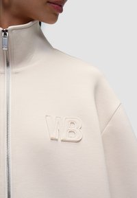 Lys beige zip-up jakke lavet af blødt stof, med høj krave og broderet "WB" logo på venstre side. Søjlebetræk i sølv.