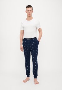 Marineblauwe sweatpants met wit Lacoste-logo patroon, elastische tailleband met logo-detail, gecombineerd met een fitted wit T-shirt, blootsvoets.