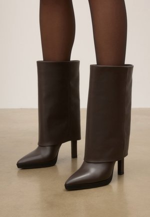 Bottes en cuir brun foncé à talons hauts avec bouts pointus portées avec des collants sombres transparents sur un sol beige lisse.