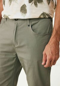 Hellgrüne Baumwollhose mit strukturiertem Oberflächen, Fronttaschen und Knopfverschluss. Der Stoff weist ein dezentes Muster auf.