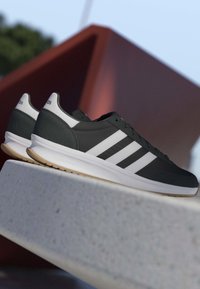 Zapatillas Adidas en blanco y negro con tres rayas apoyadas en un borde de hormigón, entorno exterior con fondo desenfocado de escultura geométrica.