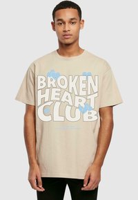 Beige bomull t-skjorte med kort erme, med iøynefallende tekst "BROKEN HEART CLUB" i hvitt og lyseblå hjerteakenter.