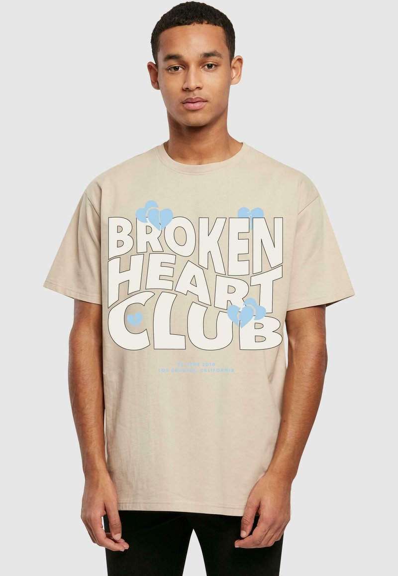 Beige bomull t-skjorte med kort erme, med iøynefallende tekst "BROKEN HEART CLUB" i hvitt og lyseblå hjerteakenter.