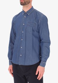 Camicia di denim con maniche lunghe, colletto a punta, taschino sul petto, colore azzurro chiaro, texture sottile e dettagli cuciti a contrasto.