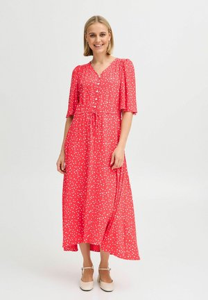 Robe maxi rouge à pois avec des manches courtes bouffantes, un décolleté en V et une taille nouée. Tissu lisse et ourlet au genou, associée à des chaussures beiges.