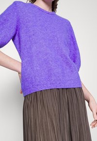 Personne portant un pull à manches courtes violet associé à une jupe plissée marron taille haute sur fond uni.