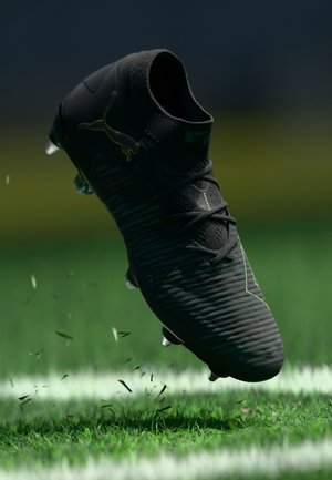 Chaussure de football noire avec une tige texturée, accents verts et lacets, suspendue au-dessus de l'herbe, montrant un mouvement dynamique avec les brins d'herbe déplacés.