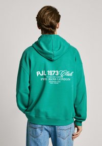 Sudadera verde con capucha hecha de un tejido suave, con texto blanco en la parte posterior: "PJL 1973 Club, Pepe Jeans London." Corte holgado, puños acanalados.