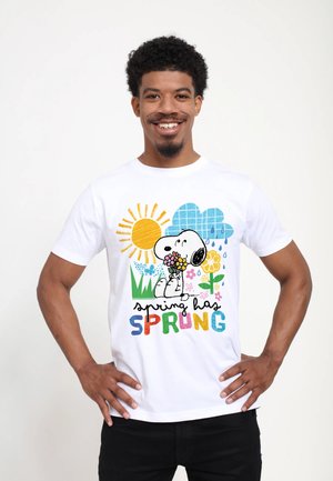 Uomo sorridente che indossa una maglietta bianca con Snoopy che tiene fiori, sole, nuvola di pioggia e testo "spring has SPRUNG" a lettere colorate.