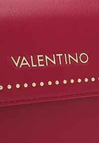 Červená kožená kabelka s pozlaceným logem "VALENTINO" a hřebíčky podél okraje. Hladký povrch s precizními detaily šití.