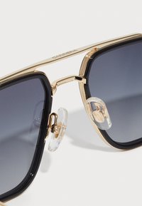 Ochelari de soare pătrați cu lentile întunecate în gradient, ramă din metal aurit și suporturi de nas transparente. Caracteristici cu margini îndrăznețe și detalii intricate pe brațe.