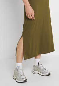 Persona che indossa un vestito verde oliva con uno spacco laterale, calze bianche fino alla caviglia e sneakers argento Adidas, in piedi su un pavimento chiaro contro un muro bianco.