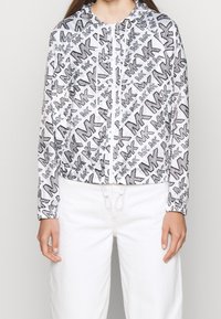 Biała krótka kurtka z czarnym wzorem graficznym z logo "MK", zamek błyskawiczny z przodu oraz regulowany ściągacz u dołu i przy rękawach.