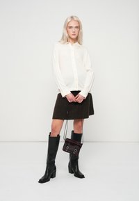 Blusa de botones ligera en color crema, falda de lana marrón, botas de cuero negro hasta la rodilla. Pequeño bolso oscuro con acentos metálicos. Diseño simple y ajustado.