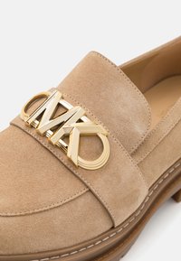 Mocassino in suede beige con una fascia larga che presenta un logo dorato. Punta rotonda e dettagli cuciti su una spessa suola marrone.