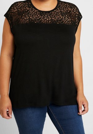 Mujer que lleva una camiseta sin mangas negra con una sección superior con un patrón de encaje y vaqueros azules sobre un fondo liso.