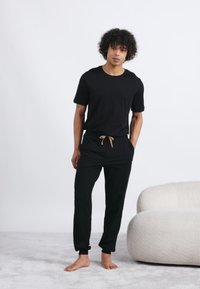 Mladic s kovrčavom kosom stoji bos, odjeven u crnu majicu i trenirke, pored bijele teksturirane fotelje na svijetlom tepihu.