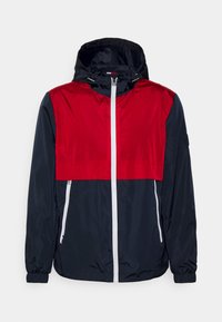 Giacca antivento leggera con la parte superiore rossa e la parte inferiore blu navy, dotata di cappuccio, cerniera bianca e due tasche laterali.
