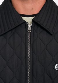 Chaqueta acolchada negra con cuello estructurado, cierre de cremallera y logotipo bordado. La tela es suave con una textura en patrón de diamante.
