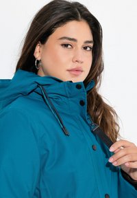 Turfärgad vattentät jacka med dragkedja, tryckknappar, justerbar huva och elastiska dragsnören, med en slät textur och synlig sömnad.