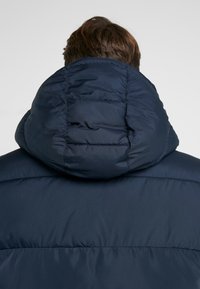 Marineblauwe pufferjas met een grote, doorgestikte capuchon. Het materiaal heeft een gladde textuur en een minimalistisch ontwerp zonder zichtbare logo's.