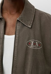 Hnědá denimová bunda se zipem, špičatým límcem a vyšitým logem "T.X. Buffalo" s červenými hvězdami na hrudi.