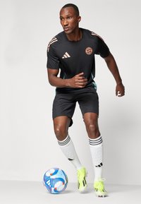 Sort atletisk t-shirt med orange detaljer, sorte shorts, hvide sokker med sorte striber og gule fodboldstøvler. En blå og rød fodbold i nærheden.