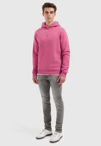 Rosa Kapuzenpullover aus Baumwollmischung, mit geripptem Saum und Bündchen, kombiniert mit hellgrauen Jeans und weißen Sneakers.