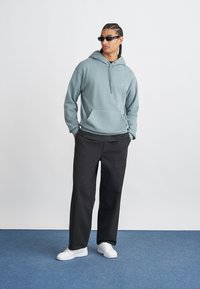 Sweatshirt com capuz em algodão verde claro com bolso frontal; combinado com calças largas pretas e ténis brancos. Modelo usa óculos de sol.