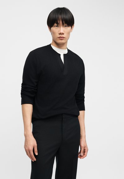 Νέος άνδρας με ίσια μαύρα μαλλιά, φορώντας μαύρο μακρυμάνικο πουκάμισο πάνω από λευκό μπλουζάκι και μαύρο παντελόνι, στέκεται μπροστά σε ένα απλό φωτεινό φόντο.