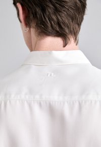 Bílá košile s límečkem viděná zezadu, s jemně vyšitým logem blízko límce, s hladkou, hedvábnou texturou.