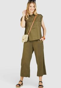 Olivgrünes, kurzärmliges Hemd mit Knopfleiste und zwei Fronttaschen, kombiniert mit passenden weiten Hosen. Die Accessoires umfassen eine geflochtene Tasche und schwarze Sandalen.
