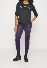 Femme portant un haut de sport noir à manches longues et un legging de sport violet avec des baskets noires, debout contre un fond blanc uniforme.