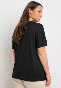 Donna con lunghi capelli castani, che indossa una maglietta nera larga e pantaloni beige chiari, in piedi contro un muro bianco texture, rivolta di lato.