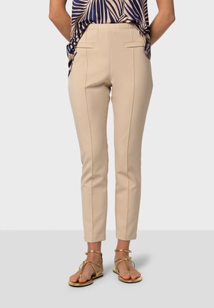 Beige op maat gemaakte broeken met smalle pijpen, zijzakken en contrasterende stiksels, gecombineerd met gouden sandalen.