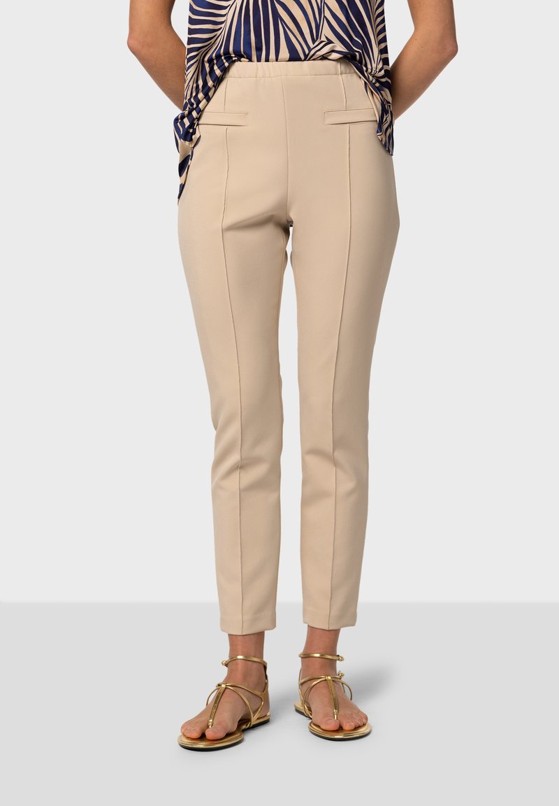 Pantalons beige sur mesure avec jambes étroites, poches latérales et détails de couture contrastants, associés à des sandales dorées.