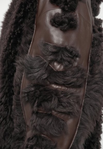 Brūnā ādas apģērbs ar greznu, teksturētu tumšu kažokādu akcentu. Ievērojama paneļu un slāņu konstrukcija ar dažādiem kažokādas garumiem.