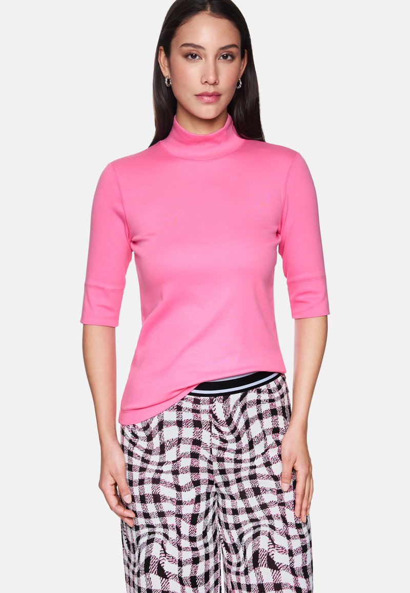 Woman wearing a bright pink turtleneck with three-quarter sleeves and black-and-white checkered pants, standing against a white background.