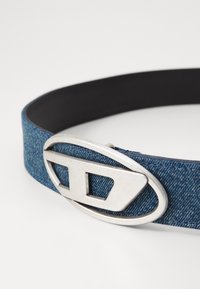 Denim belt featuring a silver oval buckle with cut-out design. The strap has a textured blue denim finish and a smooth black interior.