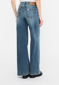 Jeans in denim a gamba larga in lavaggio blu medio, dotati di vita alta, due tasche posteriori e un'etichetta in pelle marrone sulla vita.