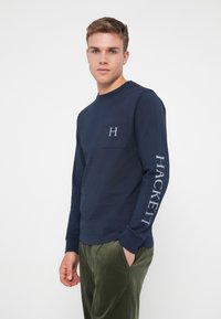 Tengerészkék, hosszú ujjú pamut póló, elöl zsebbel, rajta 'H' logó és a jobb ujján 'HACKETT' felirattal. Sima tapintású.