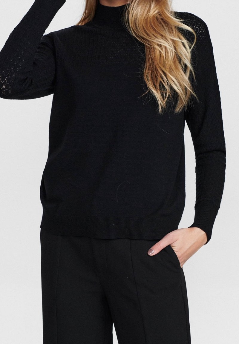 Femme portant un pull noir texturé à manches longues et un pantalon noir, avec une main dans la poche et de longs cheveux blonds sur l'épaule.