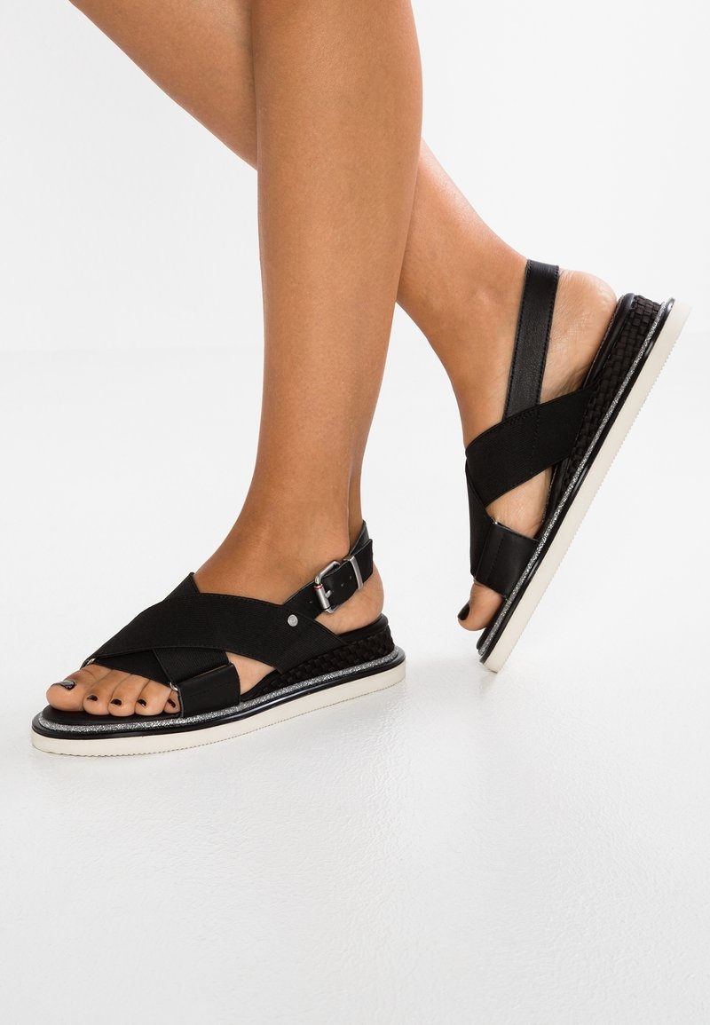 Sandales noires avec des sangles élastiques et des boucles, présentant une texture tissée sur la semelle et une semelle extérieure en caoutchouc blanc, mettant en valeur un design croisé.